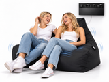 Two women laughing while reclining on a black inflatable bean bag chair. A black electronic control panel is mounted on the wall above them, with blue wave graphics indicating sound or vibration around the chair.