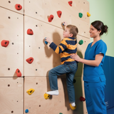 Child climbing a rock wall with a woman in blue scrubs observing.