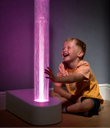 A young boy in a yellow striped shirt sits on the floor next to a tall cylindrical sensory bubble tube that glows with pink and purple light. The boy is smiling and reaching toward the light in a dimly lit room with a dark gray wall.