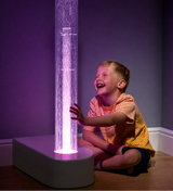 A young boy in a yellow and orange striped shirt sits on a wooden floor next to a tall, illuminated bubble tube on a white base, laughing and reaching toward the glowing water against a dark purple wall.