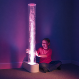A child sits on the floor next to a tall illuminated cylindrical bubble lamp, with pink and purple light glowing through the bubble tube and casting a strong colored reflection on the wall.